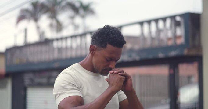 Man bowing his head, hands clasped near his face, in a moment of deep prayer and spirituality, expressing gratitude and faith, standing in a city street setting