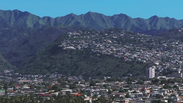 Aerial view of urban and suburban areas on Oahu Island, Hawaii, with the Ko olau mountain range in the background and a vibrant blue sky above.