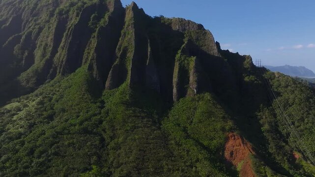 Sweeping aerial view of the Ko olau Mountain Range in Oahu, Hawaii, showcasing jagged cliffs, vibrant greenery, red soil, and visible power lines.