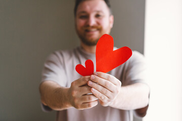 A cheerful man is showing two red paper hearts, one larger and one smaller, with a warm smile in a...