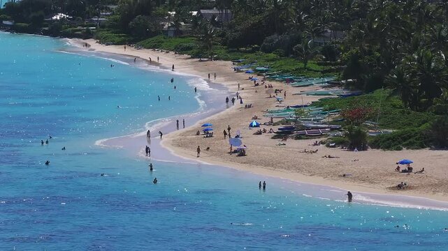 Turquoise waves lap against a sandy Lanikai beach on Oahu, Hawaii. People swim, relax under umbrellas, and walk along the shore near outrigger canoes.