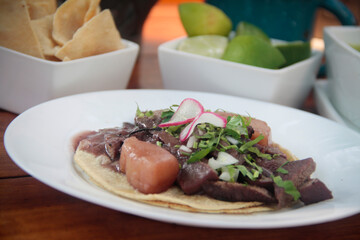 Close-up of a stewed beef tongue taco with potato, radish, and cilantro on a corn tortilla, served with chips and lemon in a rustic restaurant setting.