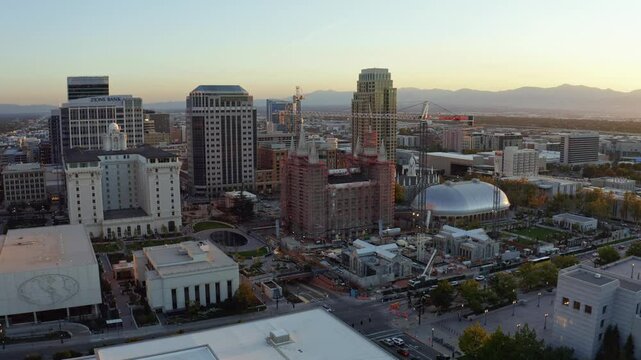 Aerial drone shot of the LDS Mormon Temple under renovation in Salt Lake City. Camera trucks right rotating around the temple amid skyscrapers, bathed in an autumn sunset with golden and orange hues