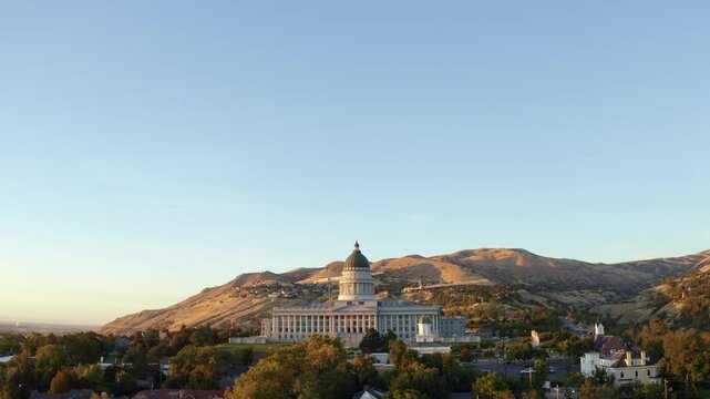 Aerial drone shot of the Utah Capitol in Salt Lake City. The camera tilts down and dollies out, revealing Capitol Hill, homes, and vibrant fall trees glowing under a golden-orange autumn sunset.