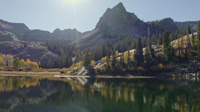Aerial drone shot of Lake Blanche in Utah&rsquo;s Big Cottonwood Canyon. The camera dollys out, showing golden aspens and green pines reflecting on an alpine lake framed by a towering mountain in autumn.