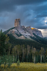 Dramatic scenic view of a towering rocky mountain peak (Chimney Rock) bathed in morning sunlight, backed by a lush forest and a dynamic cloudy sky, showcasing the beauty of nature's formations.