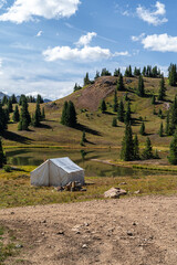 A serene outdoor campsite featuring a white canvas tent near a calm lake surrounded by hilly greenery under a bright blue sky. Ideal for outdoor enthusiasts and nature exploration activities, vertical
