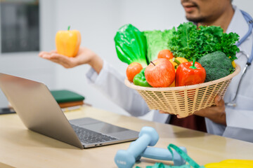 An Asian male nutritionist with a beard, wearing a lab coat, sits at a desk for an online consultation. His workspace includes fruits, vegetables, laptop, health supplements, promoting wellness.