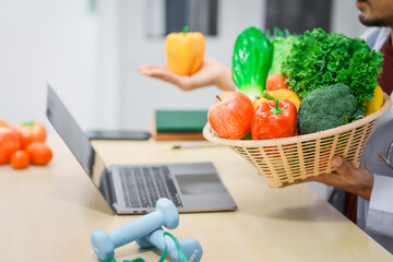 An Asian male nutritionist with a beard, wearing a lab coat, sits at a desk for an online consultation. His workspace includes fruits, vegetables, laptop, health supplements, promoting wellness.
