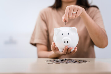 A young Asian woman in a light brown t-shirt, sitting with a piggy bank, coin, calculator, mobile, and pen. She focuses on financial stability, saving for emergencies, education, and future goals.