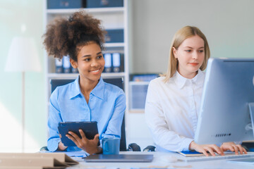 Young African American woman and Russian woman work together in business office, consulting on financial charts using desktop PC and tablet. They discuss strategies and collaborate as colleagues.