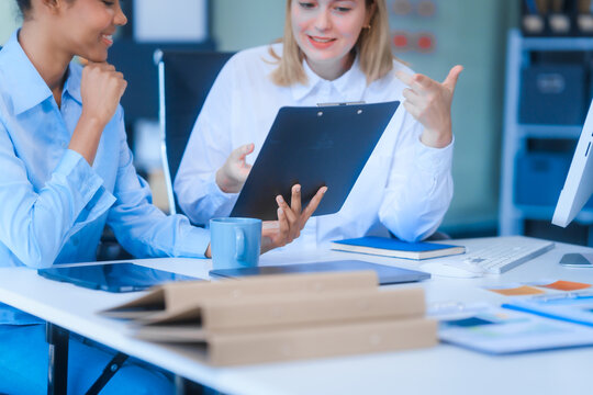 Young African American woman and Russian woman work together in business office, consulting on financial charts using desktop PC and tablet. They discuss strategies and collaborate as colleagues.