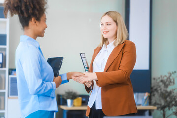 Obraz premium Two young professional women, an African American and a Russian, wearing formal suits, collaborate in a business office. They're consulting financial charts and graphs using desktop PCs, tablets