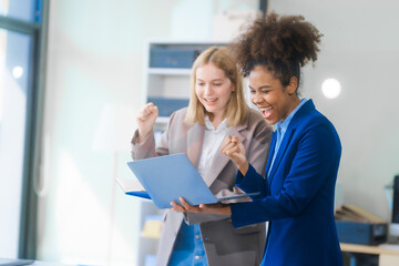 Two young women, an African American and a Russian, both wearing formal suits, collaborate in a modern business office, consulting financial charts,  engaging in professional discussions.