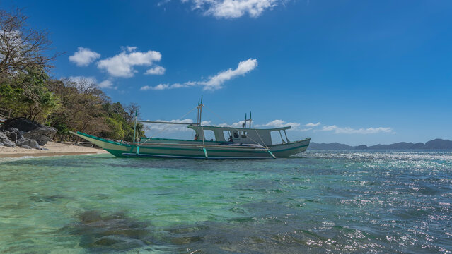 A beautiful traditional Filipino bangka boat is moored at the shore of the turquoise ocean. Picturesque boulders, green vegetation on a sandy beach. Blue sky, clouds. Philippines. Palawan. Ipil Beach 