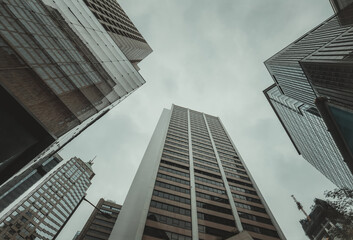 Fototapeta premium Low-angle view of towering skyscrapers in Hong Kong, reaching towards an overcast sky. The buildings are various shades of grey and brown.