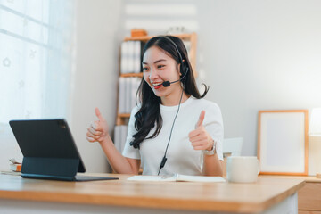cheerful woman wearing headset gives thumbs up while working on tablet at desk in bright home office