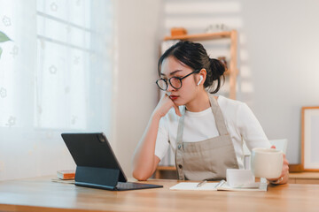 woman wearing glasses and apron works on tablet while holding coffee cup in cozy home office