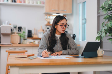 woman working at wooden table in cozy kitchen, using tablet and writing notes