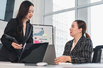 Two businesswomen discussing project in modern office with laptop and charts in background