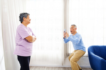 Fototapeta premium A senior couple shares a joyful moment at home as the husband playfully takes a photo of his wife with a smartphone. Bright room, happy expressions highlight love, companionship, and fun in retirement