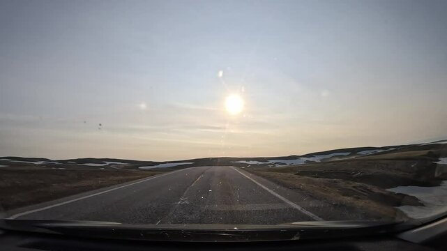 Driving through the tundra in northern Norway on the E69 road. The landscape features plains and hills covered with moss and lichen, with patches of snow. The sun shines directly ahead.