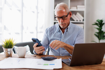 Smiling mature businessman holding smartphone sitting workplace office. Middle aged manager ceo using cell phone mobile apps and laptop. technology and solutions for business development