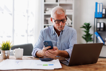 Smiling mature businessman holding smartphone sitting workplace office. Middle aged manager ceo using cell phone mobile apps and laptop. technology and solutions for business development