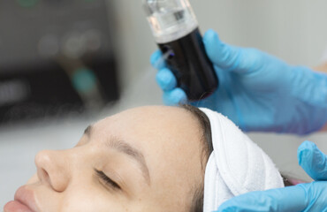 Fototapeta premium woman is getting her face done by a beautician. The beautician is holding a bottle of black liquid and a device. 