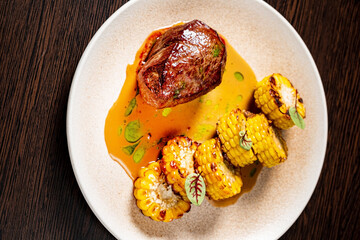 A beautifully plated dish featuring a juicy beef filet drizzled with a savory sauce, accompanied by golden grilled corn. The vibrant colors and textures invite indulgence. 