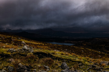 A vast landscape features a serene lake surrounded by majestic mountains, shrouded in dark clouds and mossy terrain, capturing a tranquil yet moody natural atmosphere.
