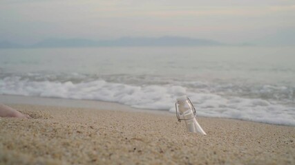 A barefoot woman discovers a drifting bottle with a rolled-up message on the sandy beach. She picks it up as gentle waves touch her feet, evoking a sense of mystery and connection.