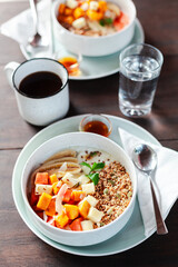 A healthy granola bowl with yogurt, mango, papaya, banana, honey, coconut flakes, and flax seeds. Served with a glass of water and a cup of coffee, creating a perfect balanced breakfast