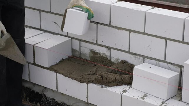 A closeup of a bricklayer worker installing white blocks and caulking brick masonry joints exterior wall with trowel putty knife outdoor. Bricklayer, masonry brick trowel in hand, mortar, brick.
