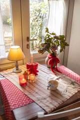 Cozy Rustic Table with Candles aA charming rustic table by the window, decorated with candles, a red bow, and a greenery arrangement. Warm natural light enhances the cozy ambiance, evoking holiday