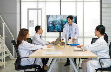 Young medical professional standing and presenting, holding a model of human skull and referencing image of brain scan displayed on a screen. The audience listening attentively with the presentation.