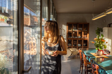 A woman in a minimalist dress holds a cup of coffee, standing by large glass windows bathed in warm sunlight. The serene atmosphere reflects slow living, relaxation, and mindful morning moments
