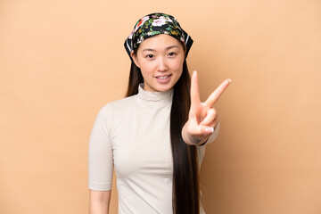 Young Chinese woman isolated on beige background smiling and showing victory sign