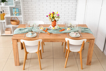 Beautiful table setting for Easter decorated with bouquet of carrots and flowers near white brick wall