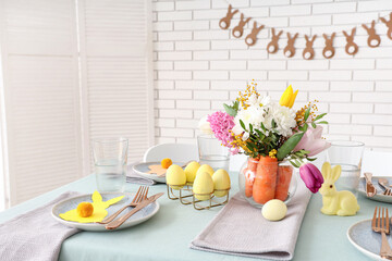 Beautiful table setting for Easter decorated with bouquet of carrots and flowers near white brick wall