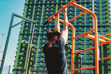 A teenager works out on the outdoor sports ground. He performs pull-ups hanging on the horizontal bar.