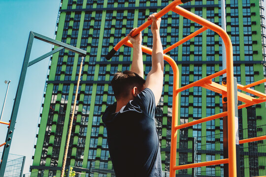 A teenager works out on the outdoor sports ground. He performs pull-ups hanging on the horizontal bar. - Powered by Adobe