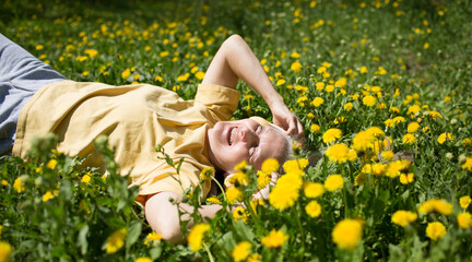  World Dandelion Day. lifestyle portrait smiling senior woman  with gray hair in headphones relaxes lying on the grass with dandelions in spring. Audio healing. meditation.  copy space. 