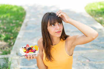 Young woman holding a bowl of fruit at outdoors having doubts and with confuse face expression