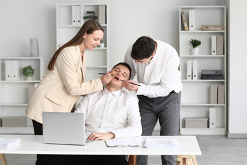 Young people drawing mustache on their sleeping colleague's face in office. April Fools' Day...