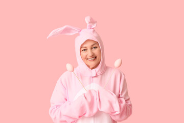 Funny senior woman in bunny costume with Easter eggs on pink background