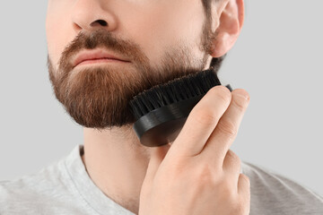 Handsome young man brushing his beard on grey background, closeup