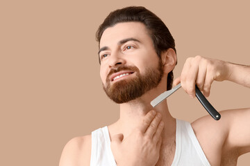 Handsome young man shaving his beard with straight razor on beige background