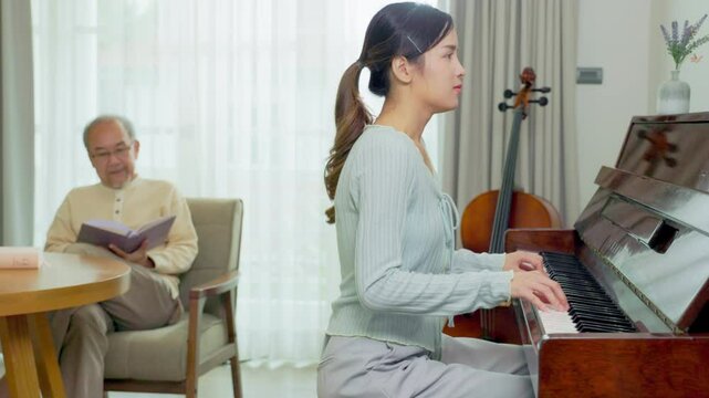 Grandfather is teaching little  daughter to play the piano at home.Portrait of happy beautiful Asian woman and her father sitting playing piano and singing together having fun and enjoying in living r