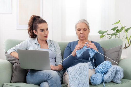 At home on the couch, a Millennials daughter uses her laptop and looking amazed to her senior mother Boomer sits nearby knitting. Contrast of two generations of different lifestyles, generation gap